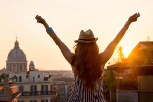 Seen from behind, a woman is standing with outstretched arms, looking out at the city of Rome at sunset in summer