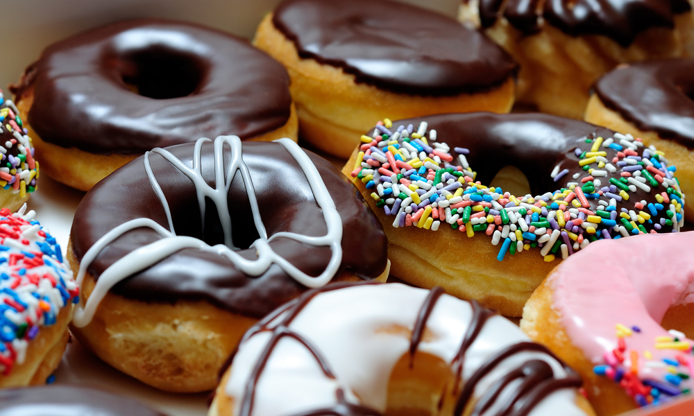 Picture of assorted donuts in a box with chocolate frosted, pink glazed and sprinkles donuts.